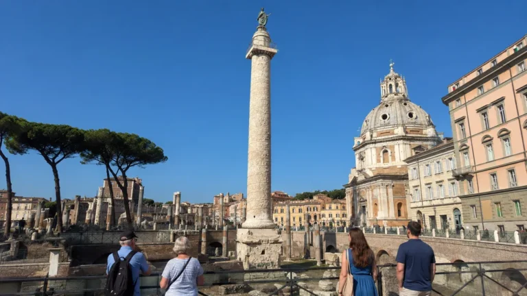 Touristes observant la colonne de Trajan à Rome avec sa frise sculptée, les ruines du forum et les monuments historiques autour.
